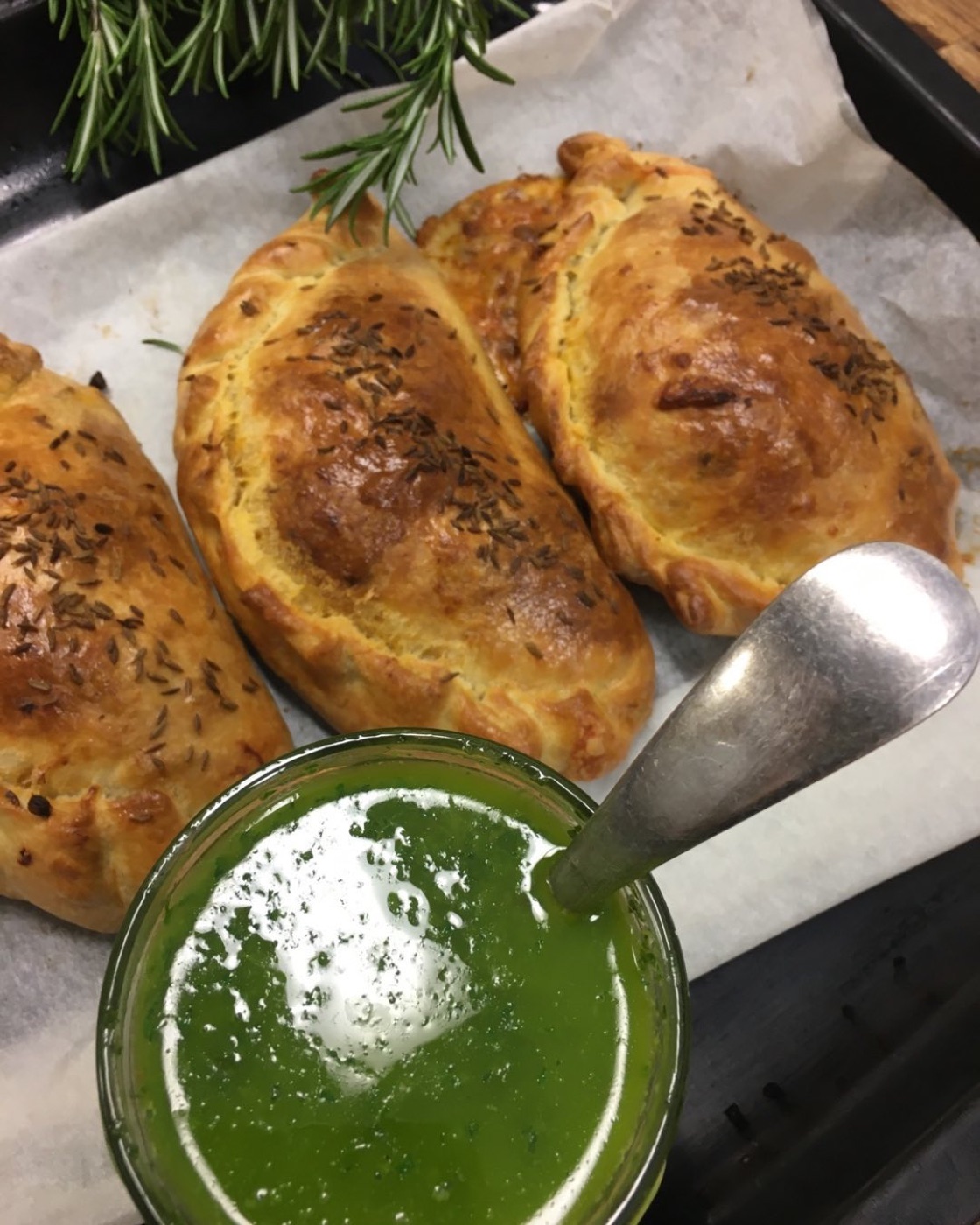 Baked pastries topped with cumin seed beside a jar of bright green sauce with a spoon