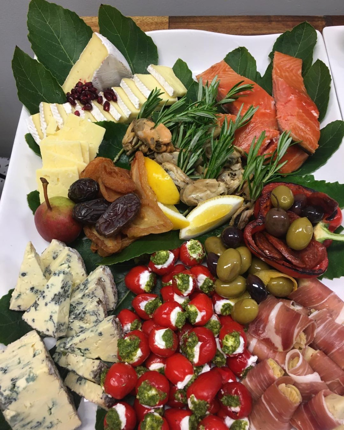 A grazing platter with cheeses, smoked salmon, olives, stuffed peppers and prosciutto on leaves